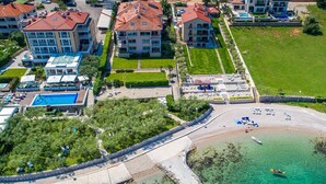 Vue aérienne d’un hôtel situé sur la côte, composé de plusieurs bâtiments, d’une piscine et d’une plage équipée de transats.