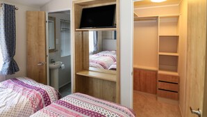 A bedroom with two beds, a TV mounted on a wooden shelf, a bathroom with a sink, and a closet with shelves. - The Goodwood Lodge (Chichester)