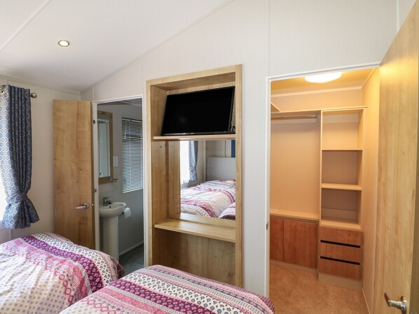 A bedroom with two beds, a TV mounted on a wooden shelf, a bathroom with a sink, and a closet with shelves. - The Goodwood Lodge (Chichester)