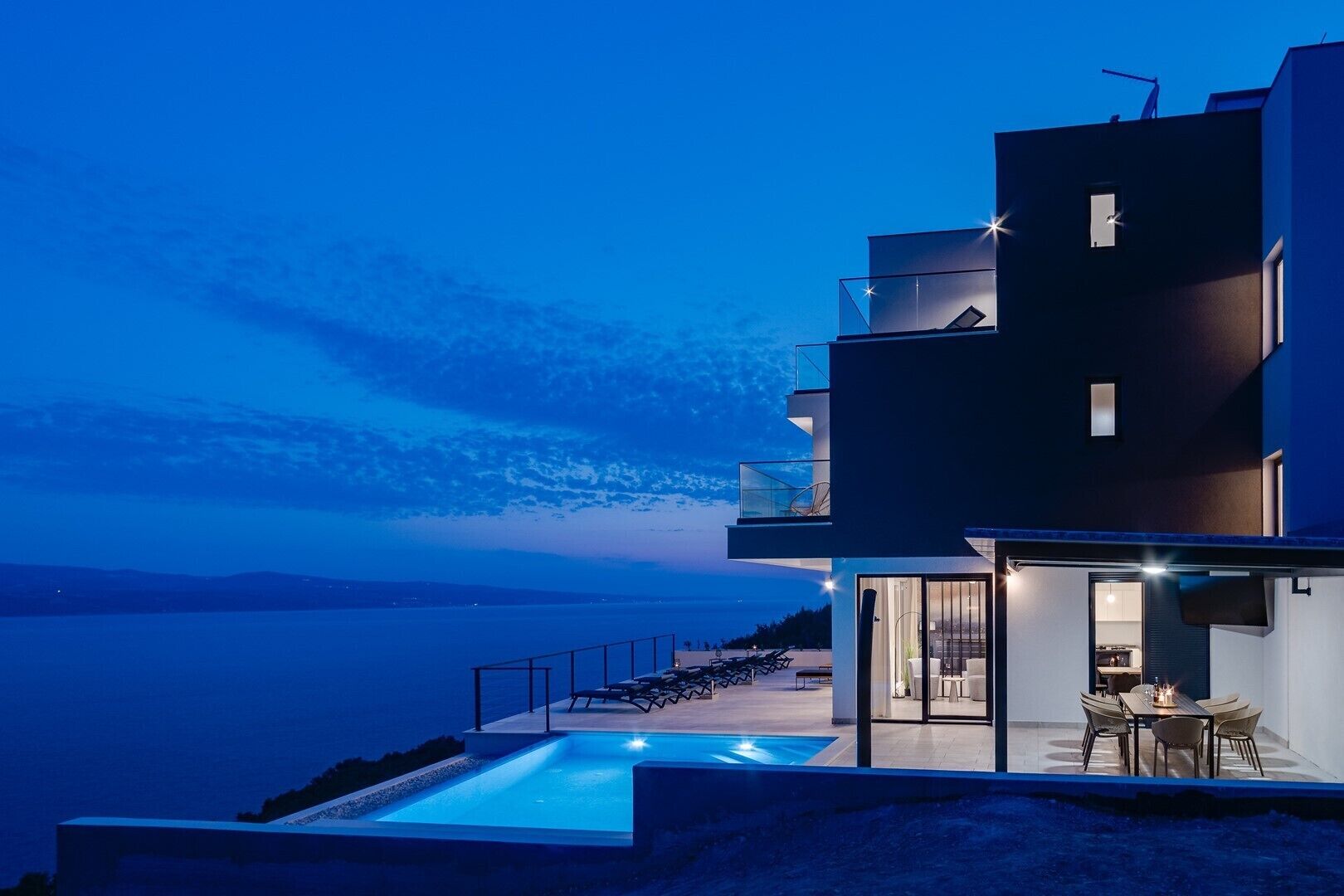 A modern multi-story building with a pool, outdoor seating, and a view of the sea.