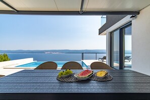 Un tavolo con vista sul mare, una piscina e una zona pranzo con sedie e un tavolo imbandito di frutta.
