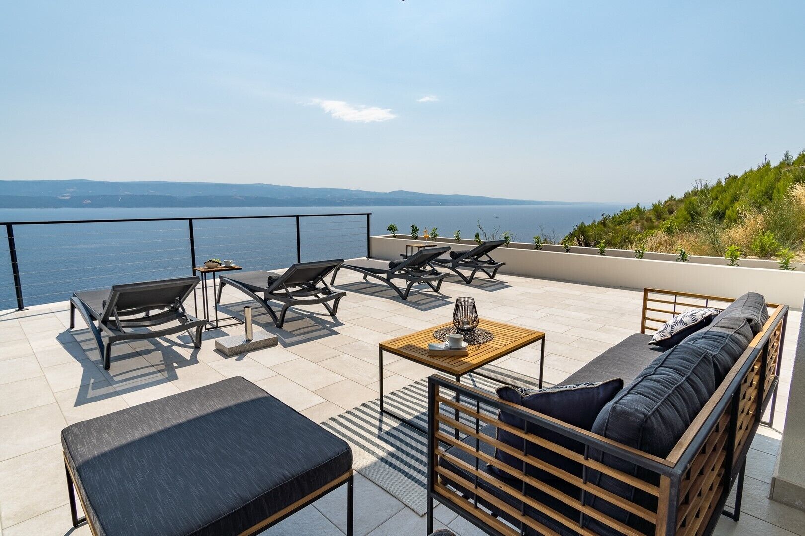 A rooftop terrace with a view of the sea, featuring a sofa, lounge chairs, and a small table.