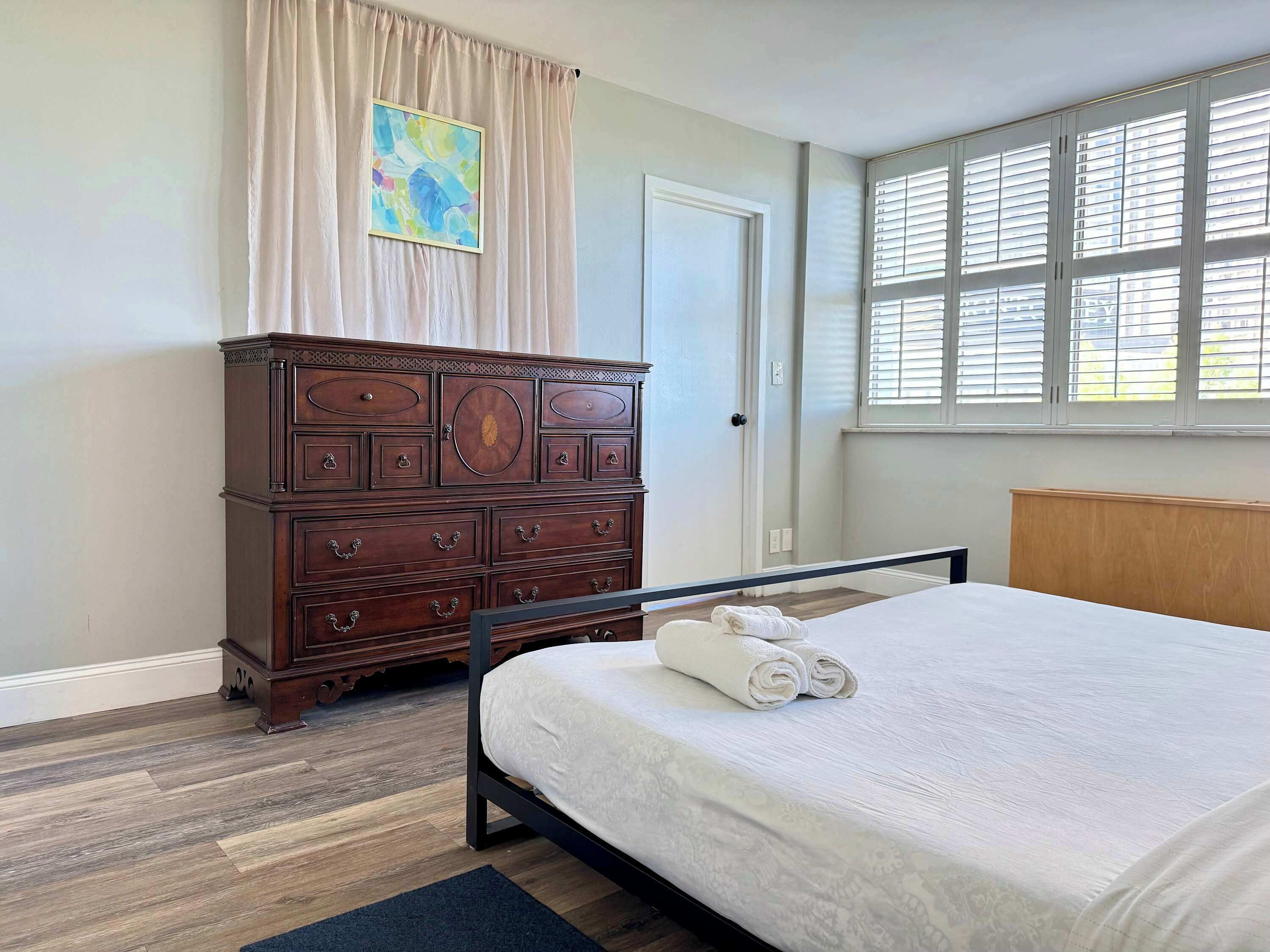 Une chambre avec une commode en bois, un lit avec des serviettes pliées, une fenêtre avec des stores et une porte.