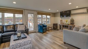 A spacious living room with a leather sofa, a grey armchair, and a blue chair. There is a wooden coffee table and a patterned ottoman. A television is mounted on the wall, and there is a window with a view of a lake and trees. - Relax in Raymond's lakefront 2-bedroom vacation home, just steps to Sebago Lake (Raymond)