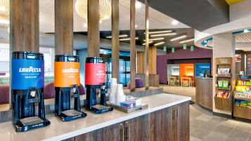 Una estación de café con tres dispensadores de café marca 'Lavazza' en diferentes colores, una barra con una máquina de café y una exhibición de vasos desechables.