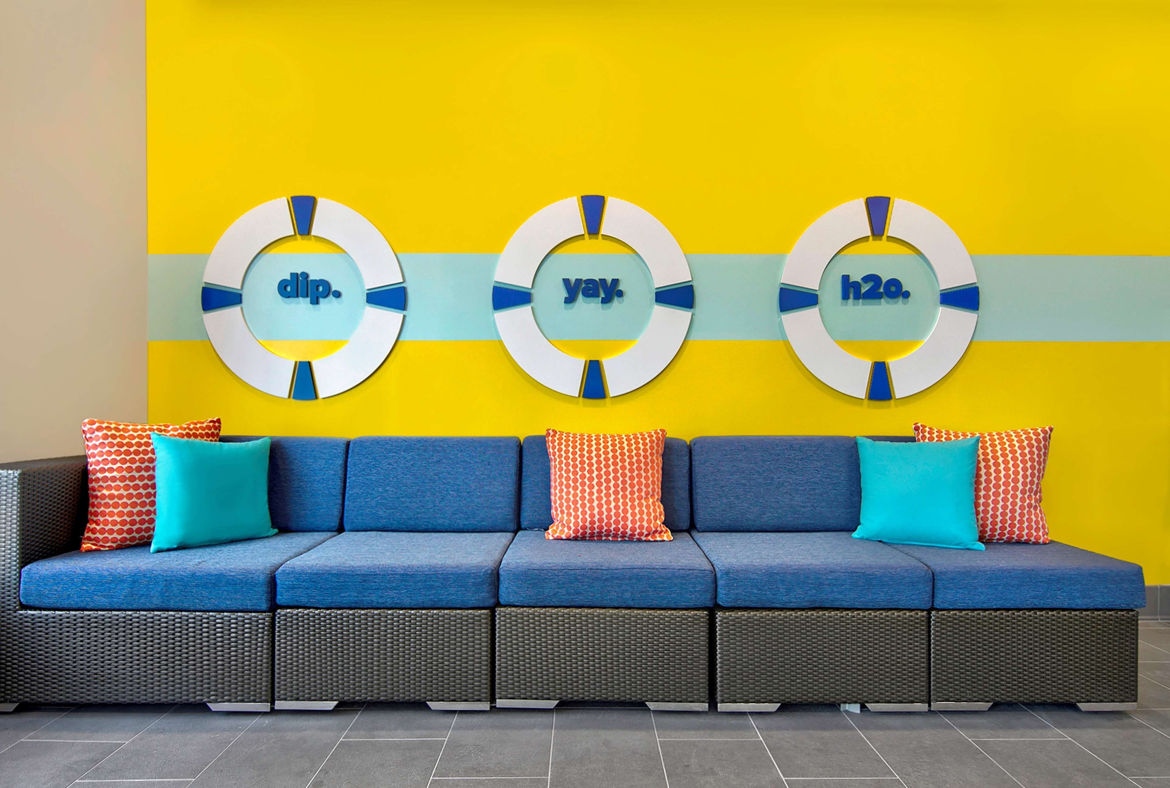 Un sofá azul con cojines estampados frente a una pared amarilla, con tres letreros circulares que dicen "dip.", "yay." y "h2o.".