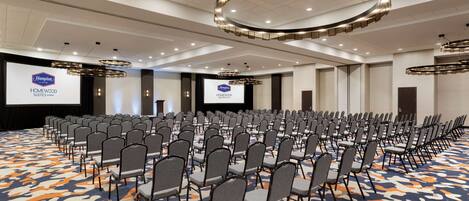 A conference room with rows of chairs, a large screen, and a logo for Hampton Homewood Suites.