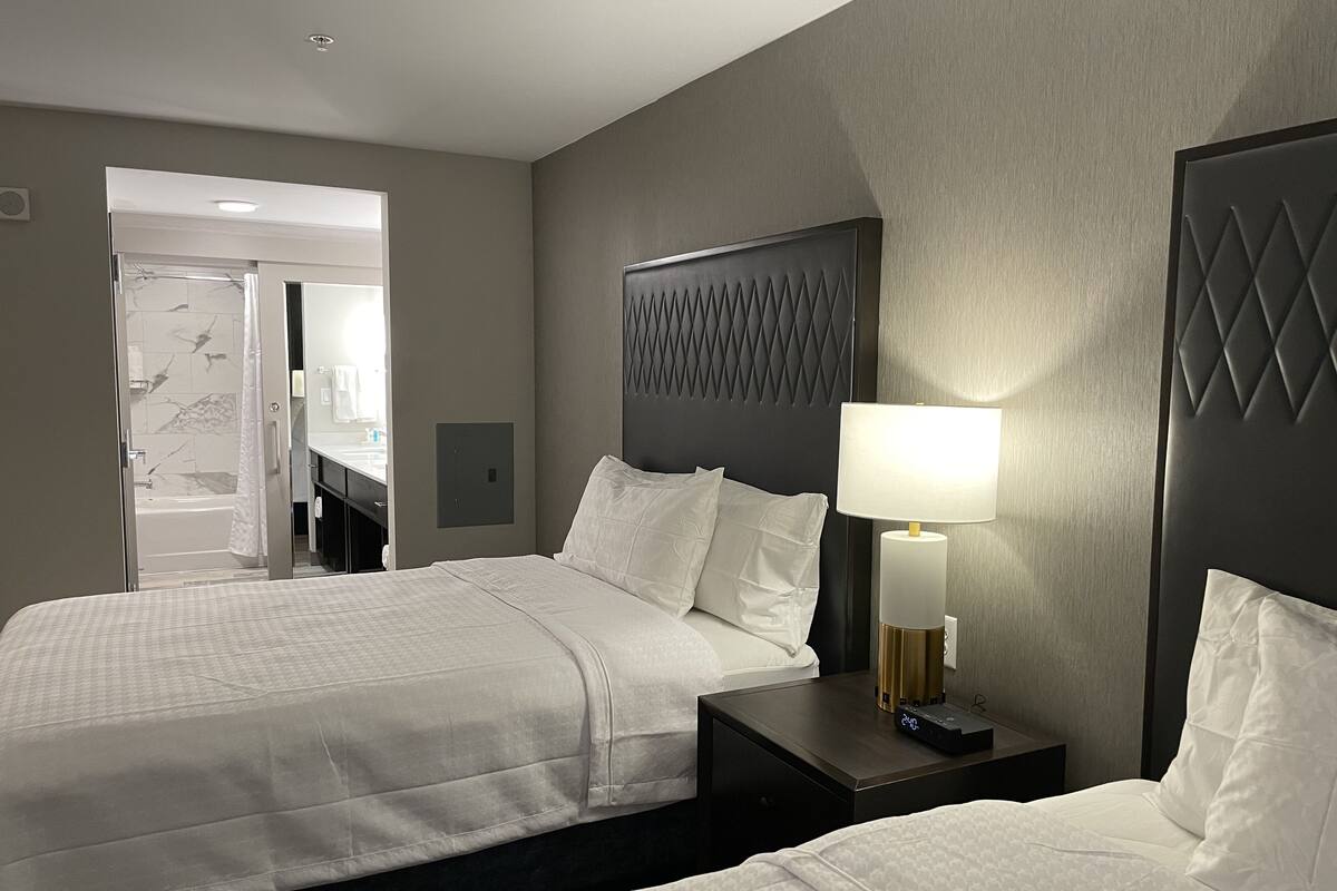 Une chambre d’hôtel avec deux lits, une table de chevet avec une lampe, une tête de lit ornée d’un motif géométrique et un espace douche visible à travers une porte ouverte.