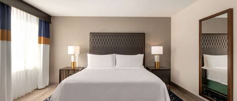 A hotel room with a large bed, two bedside lamps, a nightstand, a mirror, and a window with curtains.