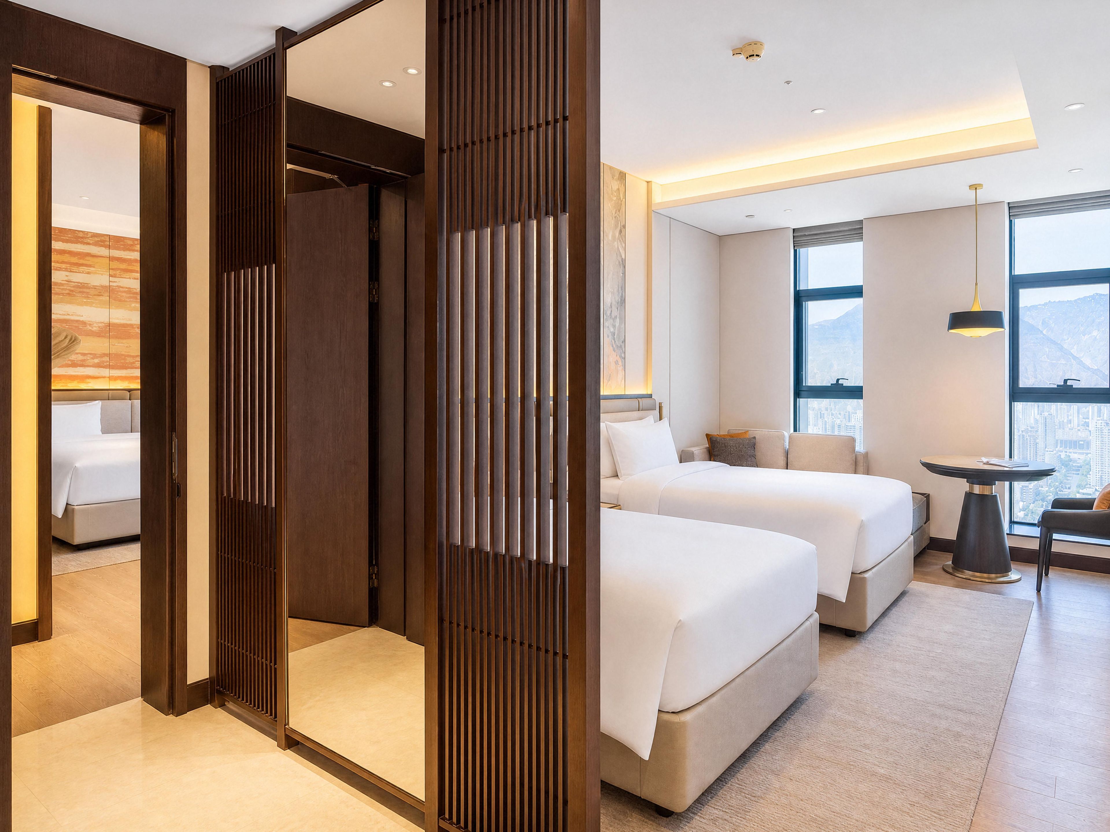 A hotel room with two beds, a sitting area with a table and chair, and large windows offering a city view.