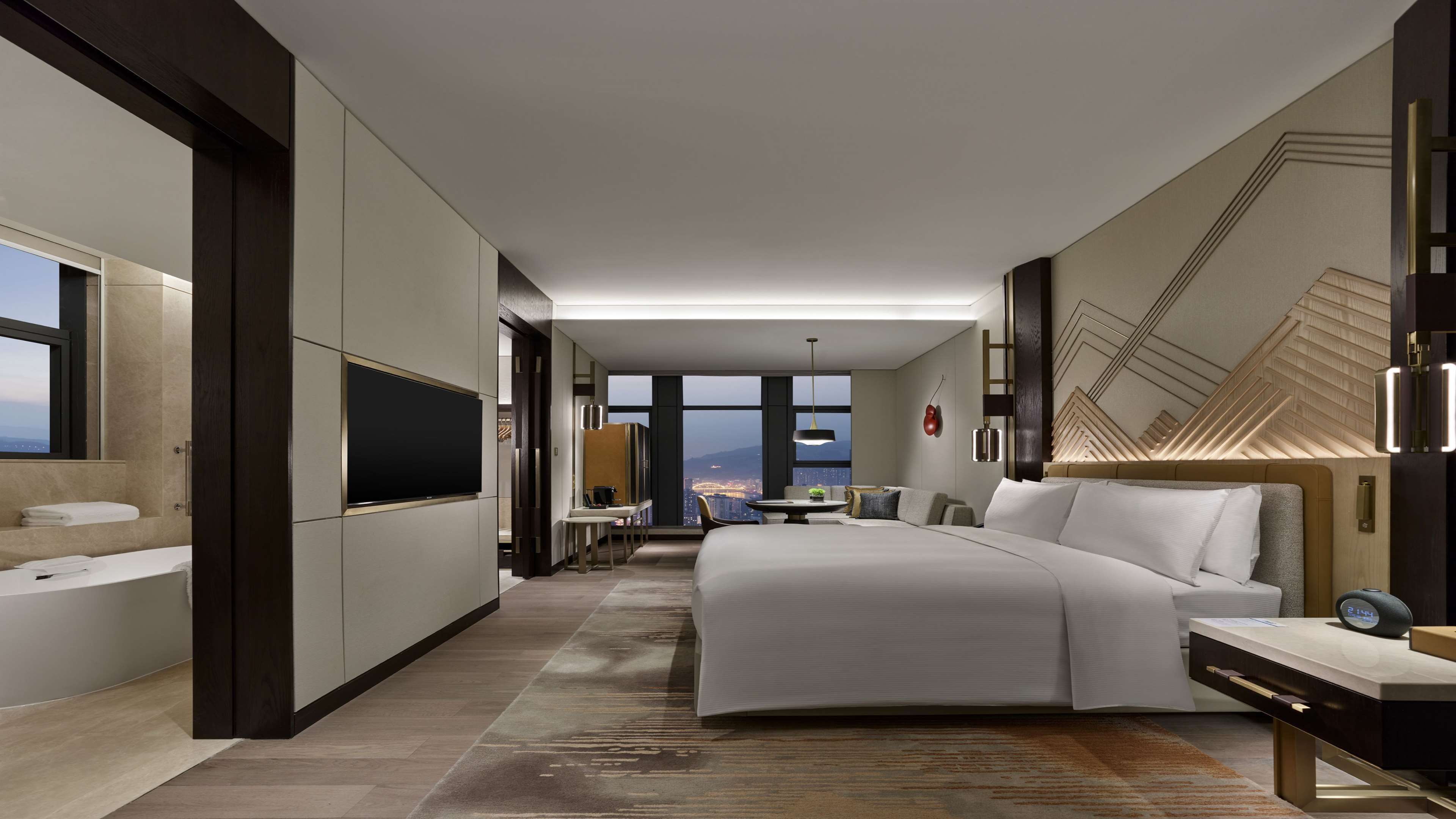 A modern hotel room with a large bed, a TV, a desk, and a view of the cityscape.