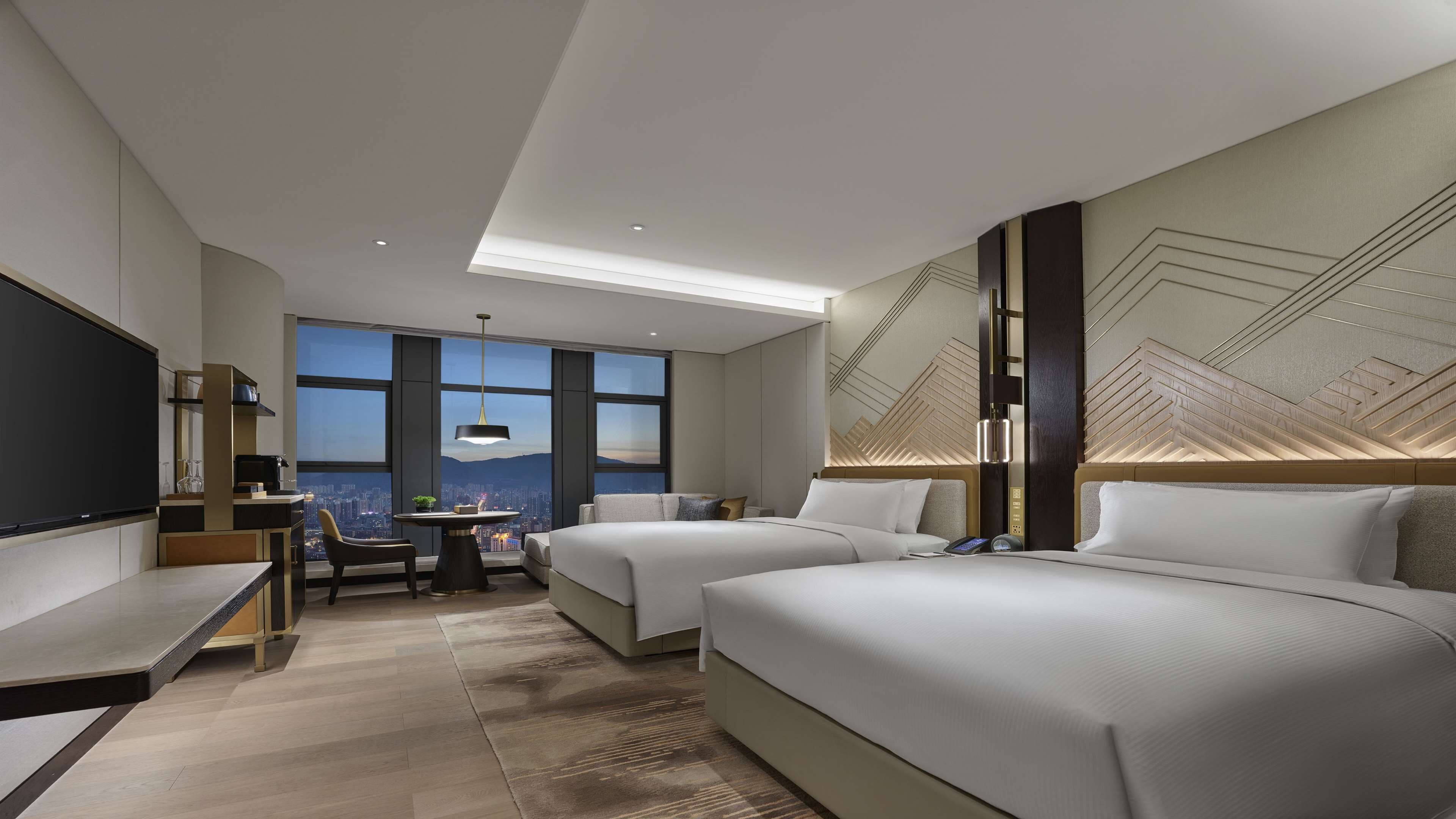 Une chambre d’hôtel moderne avec deux lits, un bureau et une grande fenêtre donnant sur la ville.
