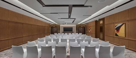 A conference room with rows of white chairs, a large screen at the front, and wooden paneling on the walls.