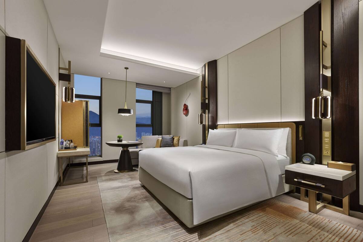 Une chambre d’hôtel moderne dotée d’un grand lit, d’une table de chevet, d’un bureau avec une chaise, d’une grande fenêtre donnant sur la ville et d’un luminaire au plafond.