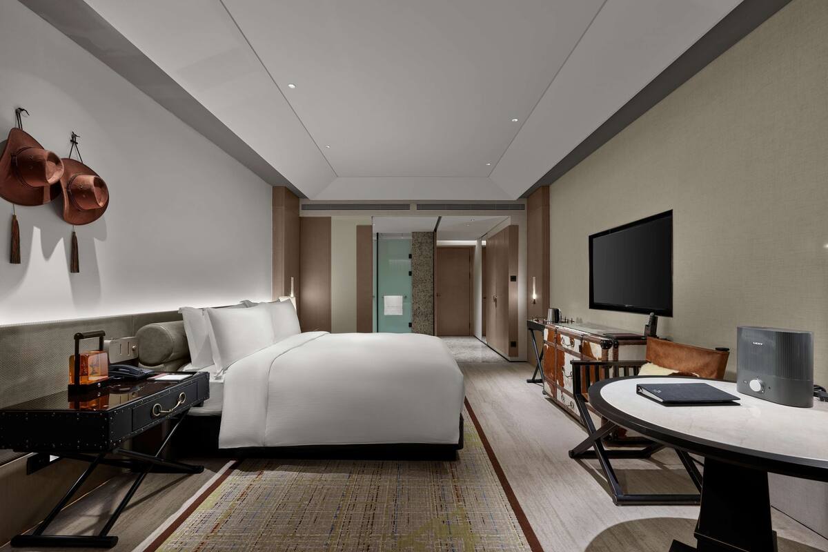 Quarto de hotel moderno com uma cama grande, uma mesa com computador, uma televisão de tela plana e um espaço de estar com mesa e cadeiras.