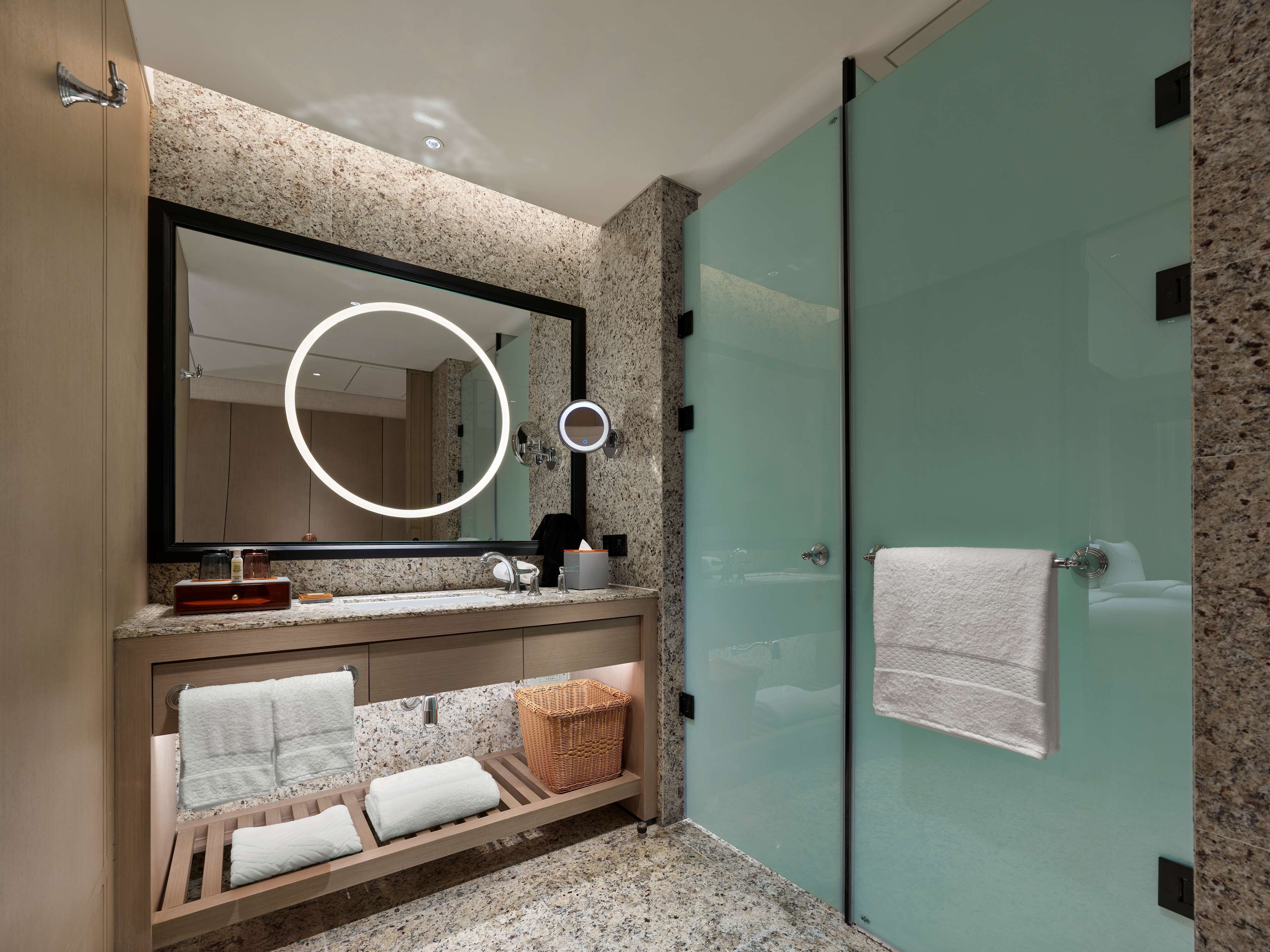 Un baño moderno con encimera de granito, un espejo grande con luz circular, cabina de ducha de vidrio y tocador de madera con toallas.