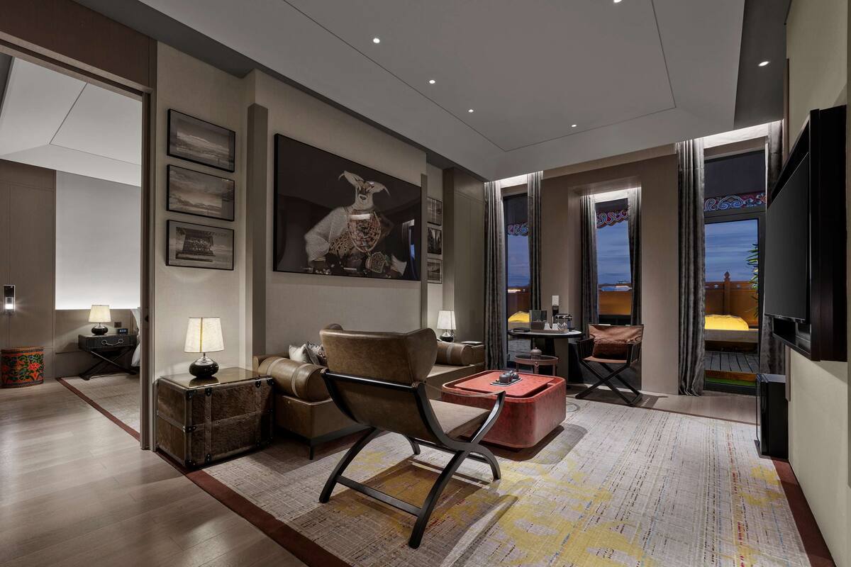 Quarto de hotel moderno com sofá, poltrona e uma pequena mesa. Há uma pintura grande na parede, uma televisão e uma janela com cortinas.