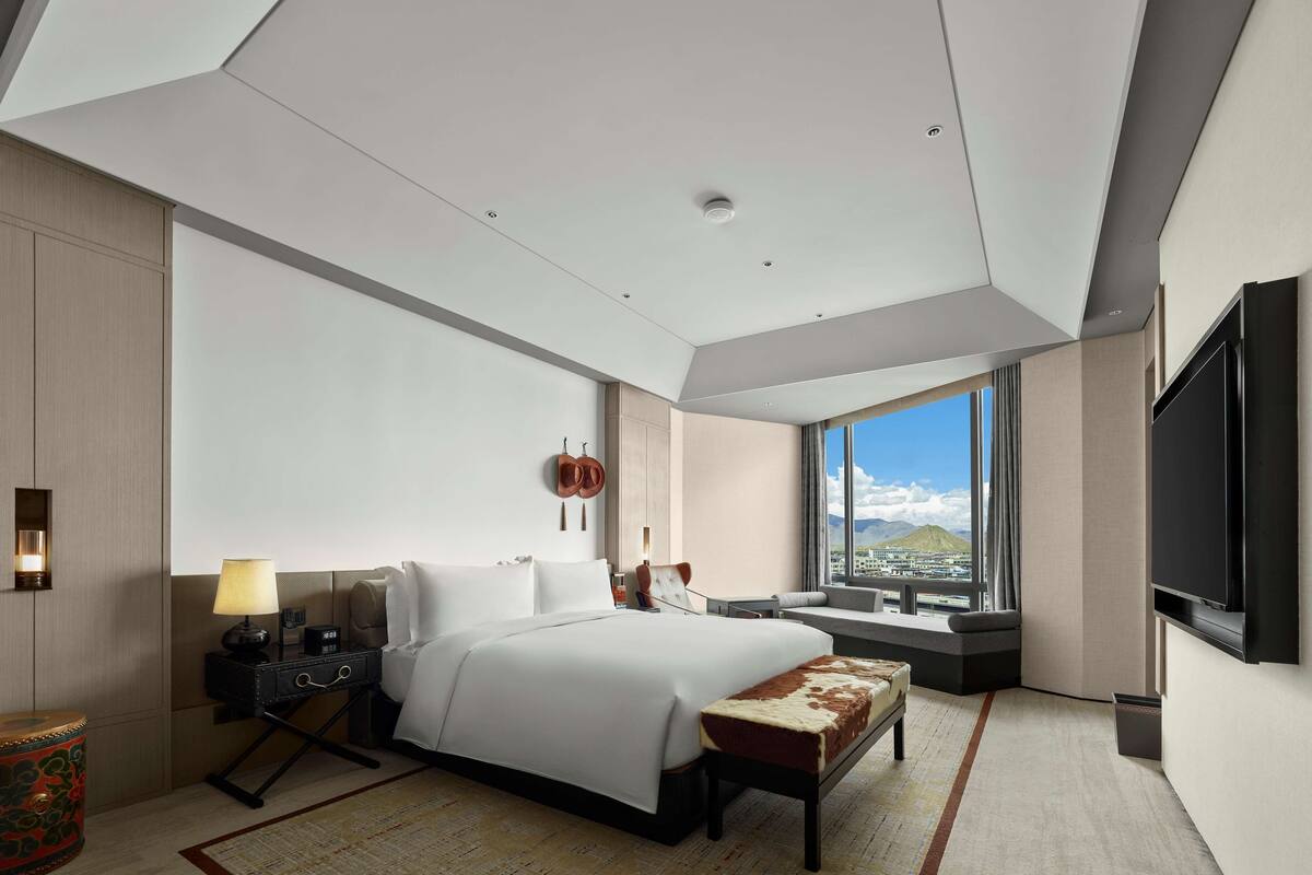 Quarto de hotel moderno com uma cama grande, uma TV de tela plana, uma escrivaninha com abajur e vista para uma montanha por uma janela ampla.