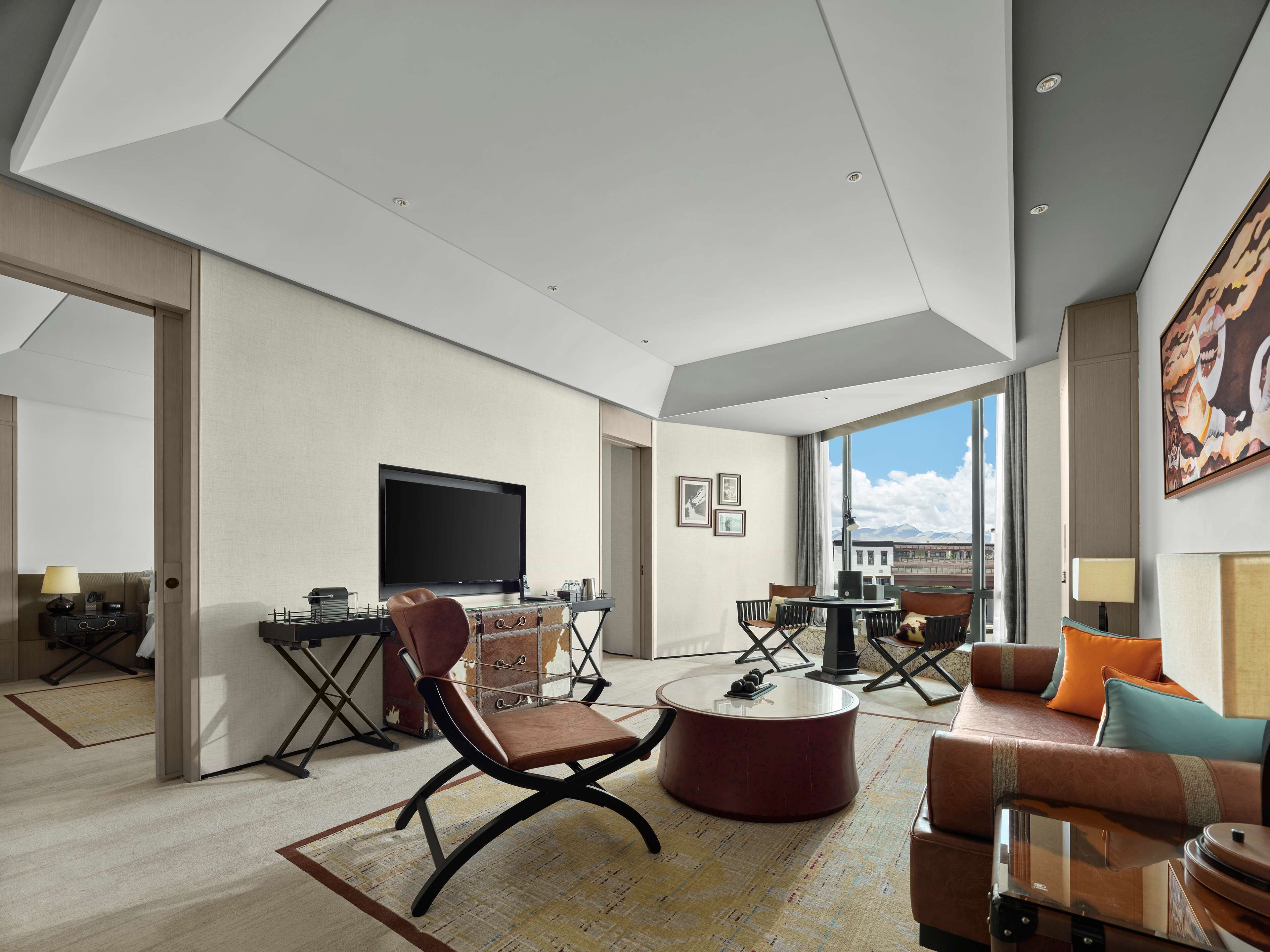 A modern hotel room with a large TV, comfortable seating, a dining area, and a view of the city.