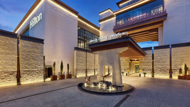 A modern hotel with a stone facade, a prominent 'Hilton' sign, and a central courtyard with a water feature and bird sculptures. - Hilton Shigatse (Shigatse)