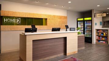 A hotel reception desk with a digital kiosk, a refrigerator stocked with beverages, and a snack bar.