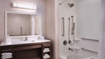 A modern bathroom with a large mirror, a sink with a towel dispenser, a vanity with a towel rack, a shower with a handheld showerhead, and a bathtub with a small table.