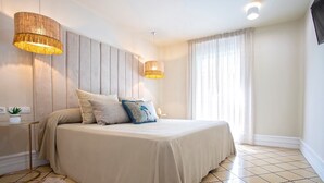 Um quarto com cama, duas luminárias pendentes, uma mesa de cabeceira com planta e uma janela grande com cortinas.