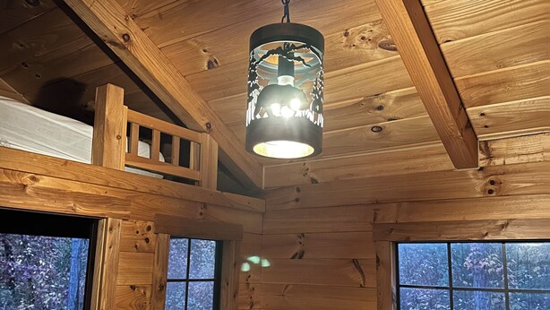A cabin interior with wooden walls, a ceiling fan, and a window with a view of a forest. - Log Cabin Sleeps four (Big Rapids)