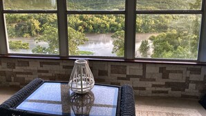 Ein Zimmer mit großem Fenster, das einen Blick auf bewaldetes Gebiet und einen Fluss bietet. Ein Glastisch trägt einen gewobenen Stuhl und eine dekorative Glasvase.