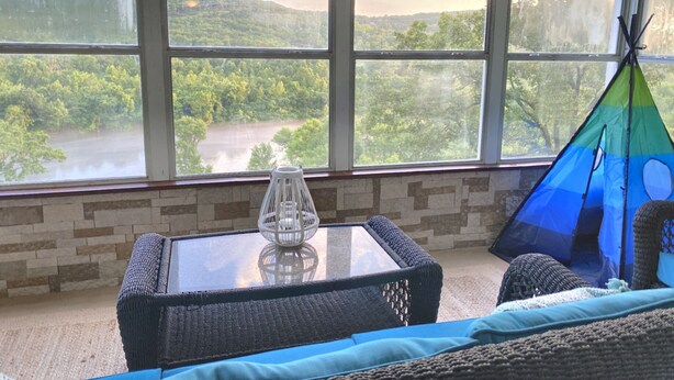 Ein Zimmer mit großem Fenster, das auf einen Fluss und Hügel blickt, einem Glastisch mit weißer Vase sowie einem blaugrünen Zelt.