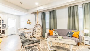A modern living room with a sofa, coffee table, and a hanging chair. There is a kitchen area with white cabinets and a dining table.