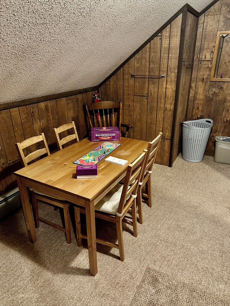Una mesa de comedor de madera con sillas, un juego de mesa y un bote de basura en una habitación con paredes de madera y un techo con textura.