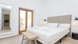 Ein Schlafzimmer mit einem Bett, Nachttischlampen, einer hölzernen Bank und Blick durch eine Tür in einen Außenbereich.