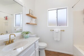 Une salle de bain avec un lavabo blanc, un robinet doré, un miroir, des toilettes et une fenêtre.