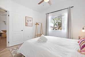 Une chambre à coucher avec un lit, une fenêtre avec des rideaux, un ventilateur de plafond et une table de chevet avec une lampe.