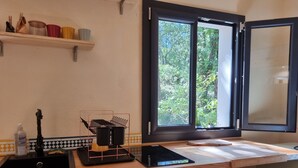 Una cocina con ventana, una repisa con recipientes de colores, un lavamanos y una encimera con hervidor y tabla de cortar.