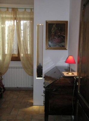 Una habitación con un piano, una lámpara roja, un cuadro en la pared y una ventana con cortinas translúcidas.