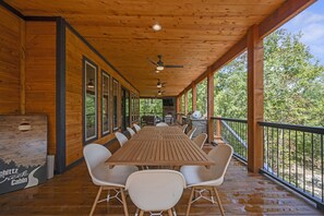 Eine überdachte Holzterrasse mit langem Tisch und weißen Stühlen, umgeben von Glastüren und -fenstern, mit Blick auf ein bewaldetes Gebiet.