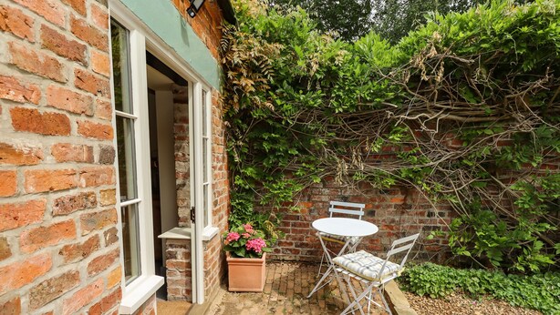 A brick house with a small patio area featuring a white table and chairs, a potted plant with pink flowers, and a brick wall with climbing plants. - The Old Mower Shed (Grantham)