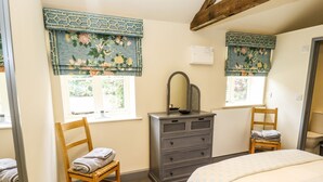 A bedroom with a bed, two wooden chairs, a dresser with a mirror, and two windows with floral curtains. - The Old Mower Shed (Grantham)