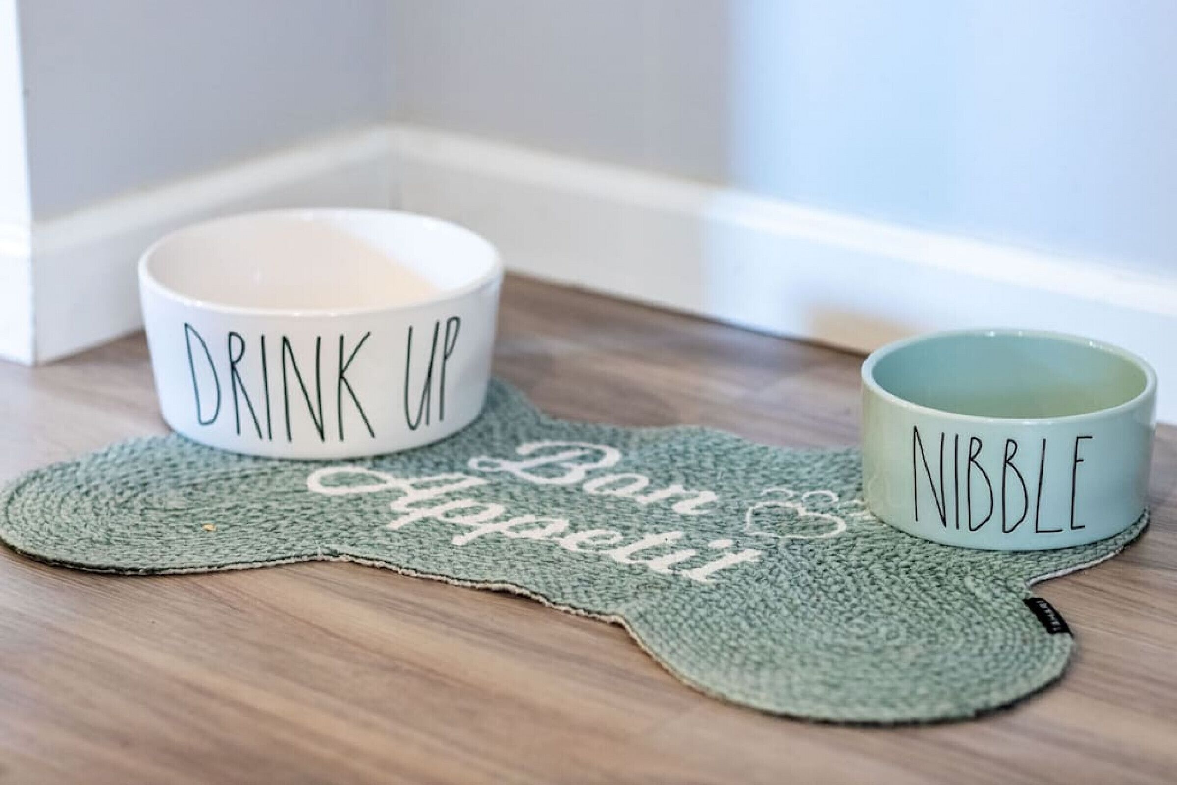 Dos tazas de cerámica sobre un mantel tejido con las palabras “Drink Up” y “Nibble”.
