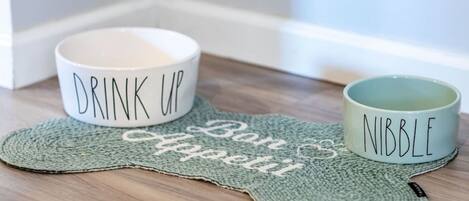 Dos tazas de cerámica sobre un mantel tejido con las palabras “Drink Up” y “Nibble”.