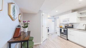 A modern kitchen with white cabinets, a built-in oven, and a microwave. There is a dining area with a wooden table and green chairs.