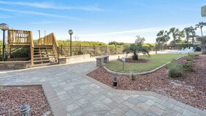 Un espace extérieur pavé comprenant une aire de jeux en bois, un banc en pierre et un jardin aménagé avec un petit étang et un éclairage décoratif.