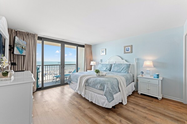 A bedroom with a large window, a bed with blue bedding, a nightstand, and a view of the beach.