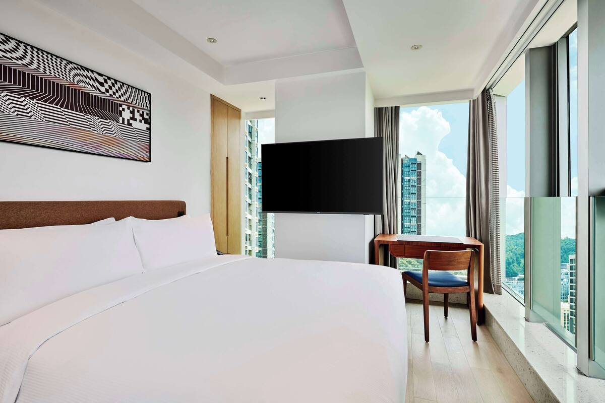 Une chambre d’hôtel dotée d’un grand lit, d’un téléviseur à écran plat fixé au mur, d’une table à manger avec des chaises et offrant une vue sur la ville grâce à de grandes fenêtres.