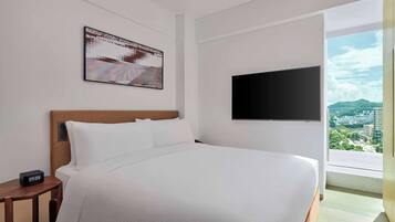 A hotel room with a large bed, a flat-screen TV mounted on the wall, a small bedside table with a clock, and a window offering a city view.
