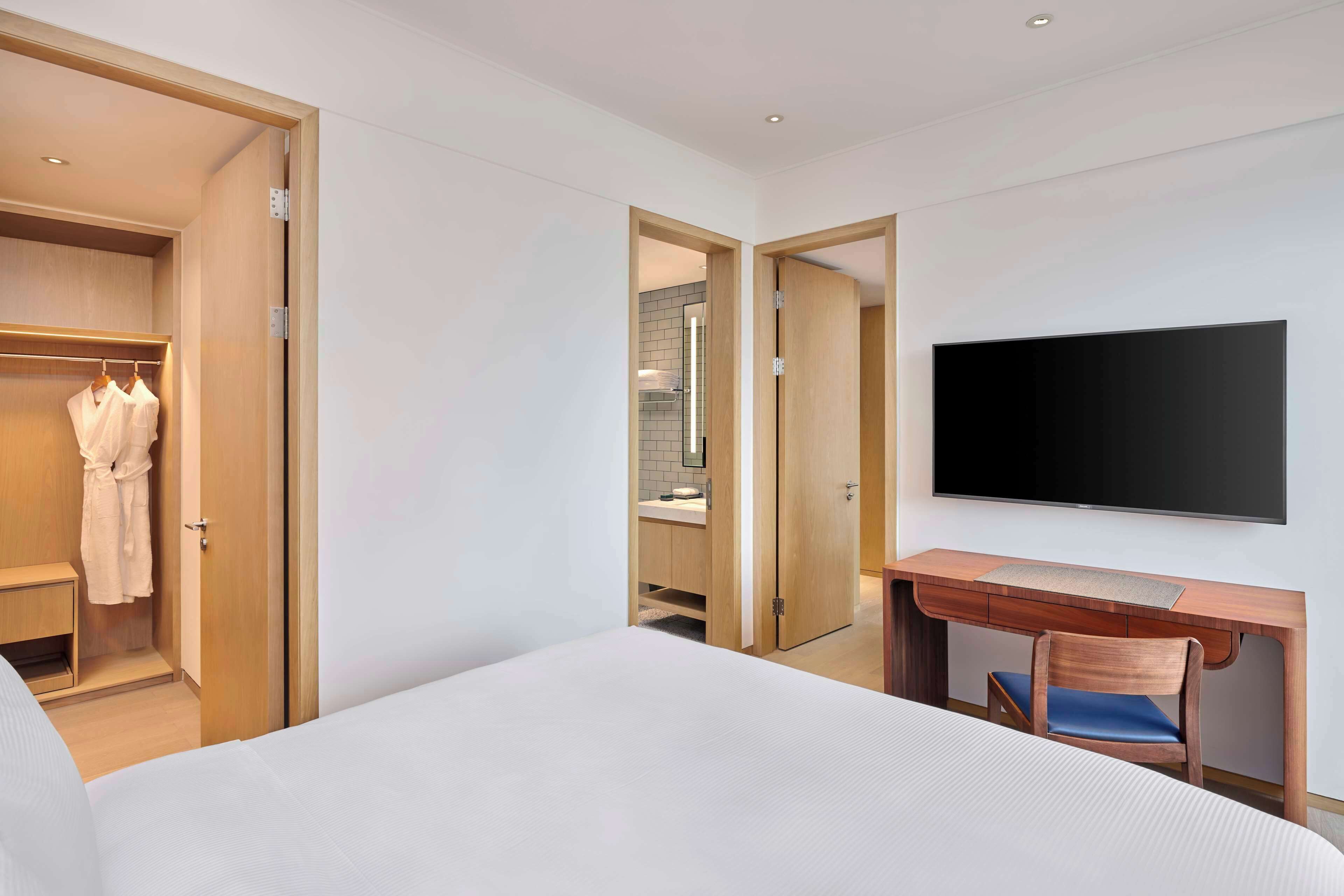 Habitación de hotel con cama, escritorio con silla, televisor, armario y baño visible a través de una puerta abierta.