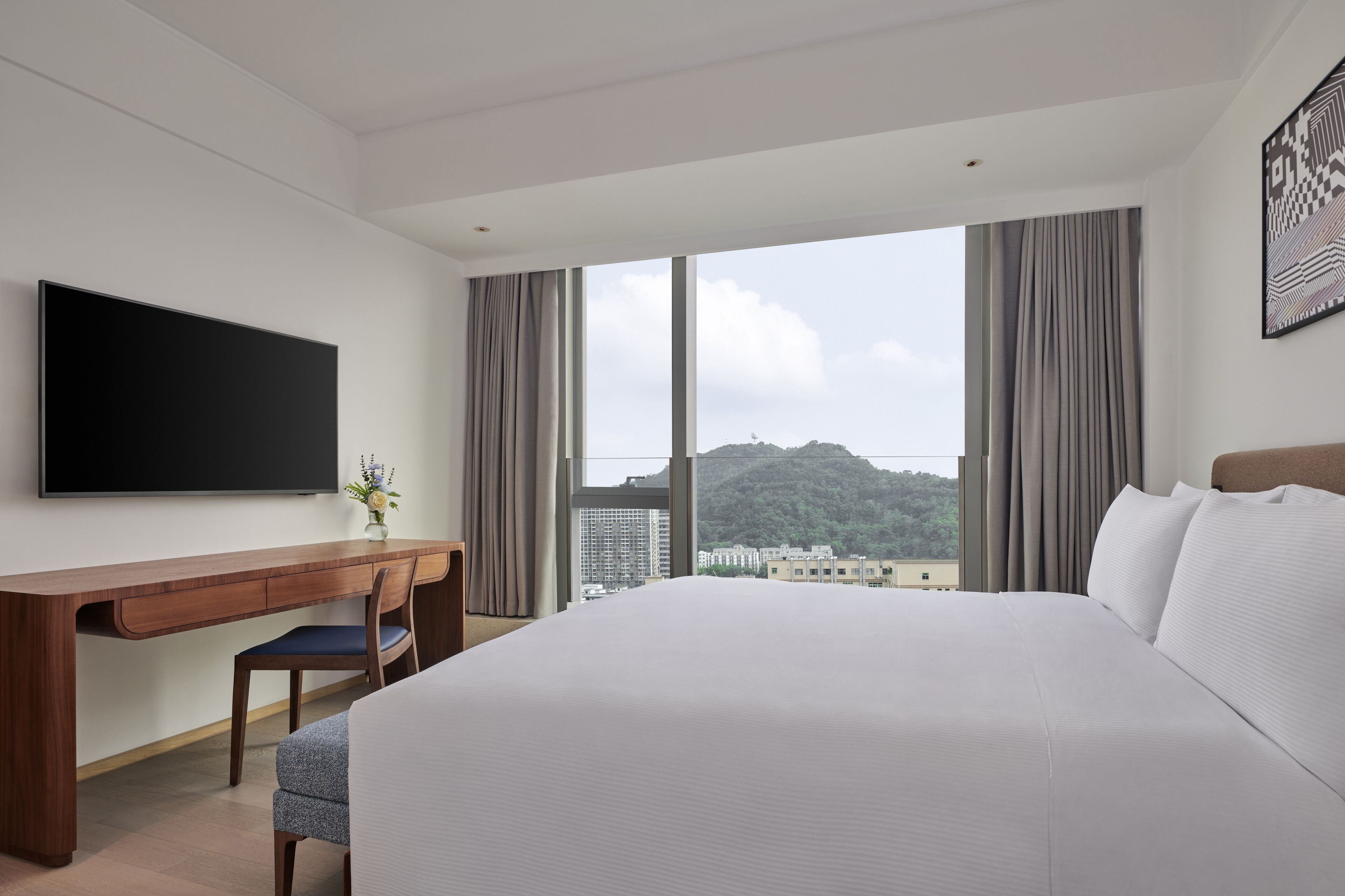 A hotel room with a large bed, a desk with a chair, a flat-screen TV, and a large window with curtains.