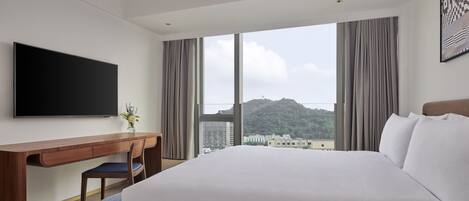 A hotel room with a large bed, a desk with a chair, a flat-screen TV, and a large window with curtains.